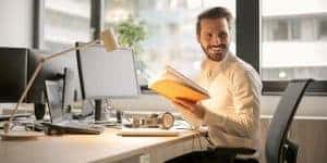 A man working on his workstation, holding a book in his hands also looking back and smiling
